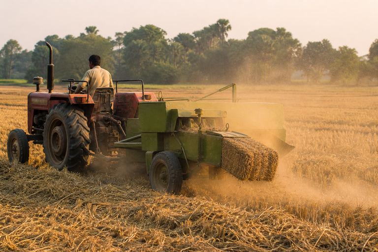 A tractor-drawn baler working across a harvested field, discharging a rectangular bale with dust in the low sunlight.